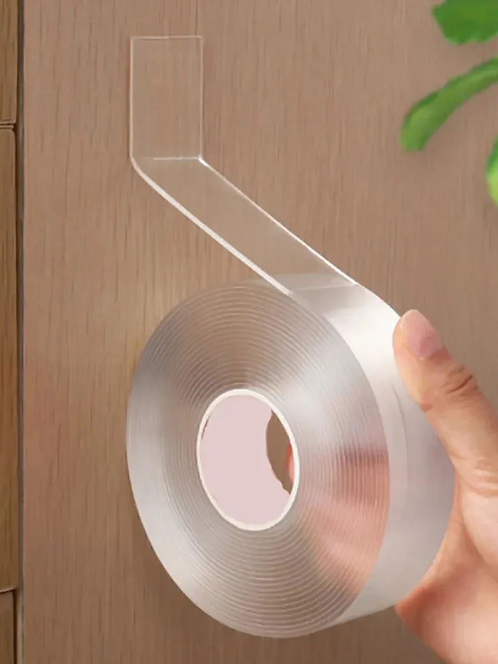 A hand is holding a roll of clear double-sided adhesive tape with a dispenser attached, against a wooden background.