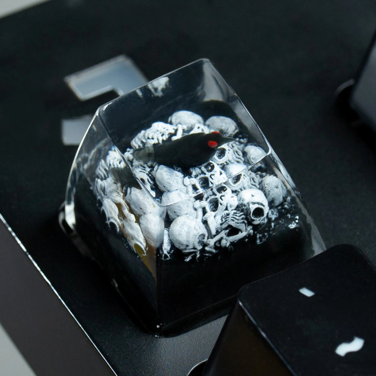 A black resin keycap featuring a gothic raven and skull design, displayed on a dark surface.