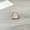 Un pequeño pin de esmalte beige claro en forma de gato adorable reposa sobre una superficie de madera.