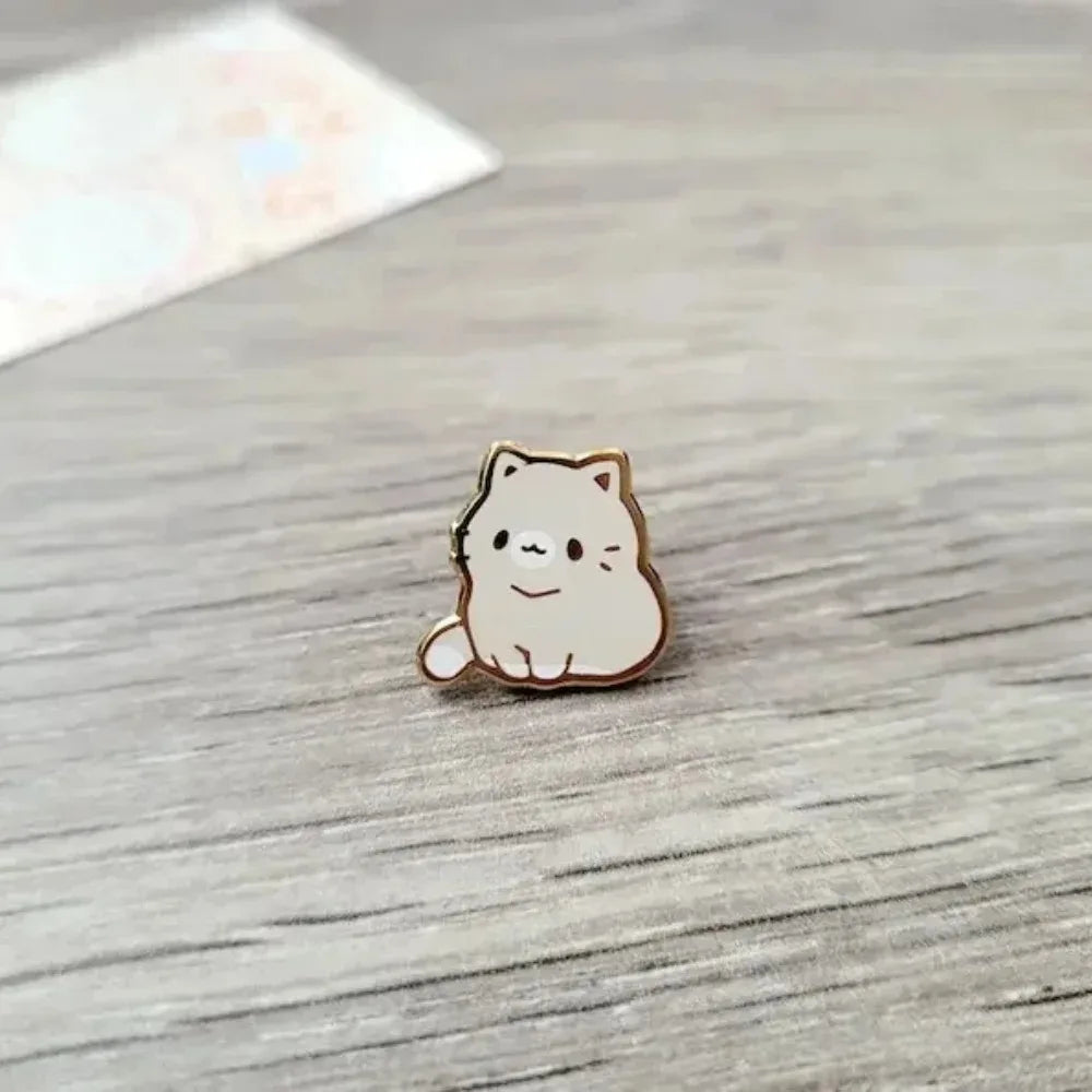 Un pequeño pin de esmalte beige claro en forma de gato adorable reposa sobre una superficie de madera.