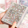 A hand holds a spiral-bound sticker book filled with cute cartoon stickers of desserts and food for scrapbooking