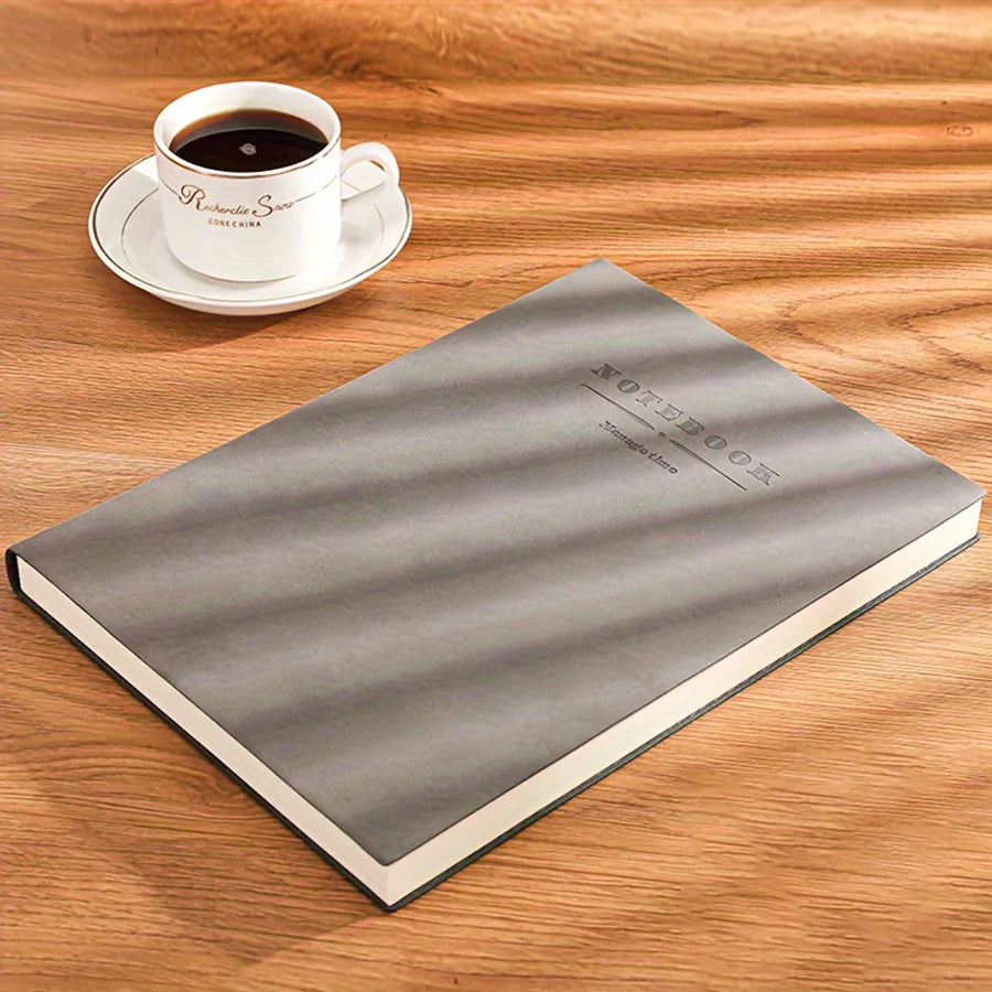 A gray notebook lies on a wooden surface next to a white cup of coffee.