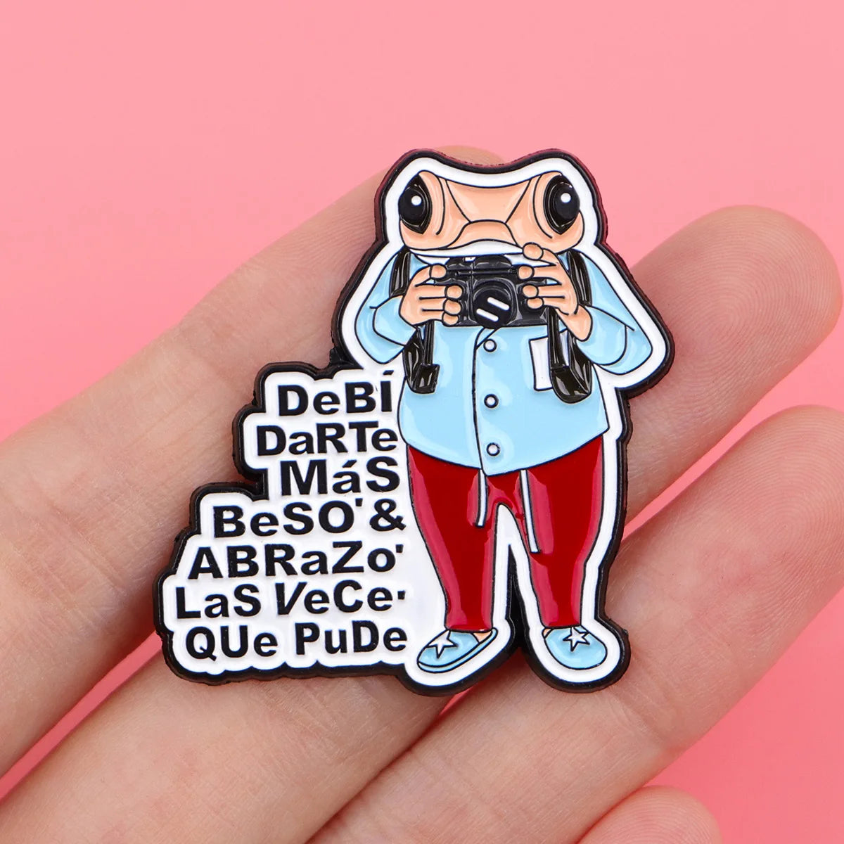 An enamel pin featuring a frog character holding a camera and text in Spanish