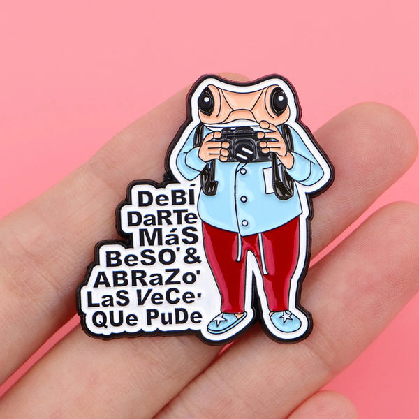 An enamel pin featuring a frog character holding a camera and text in Spanish