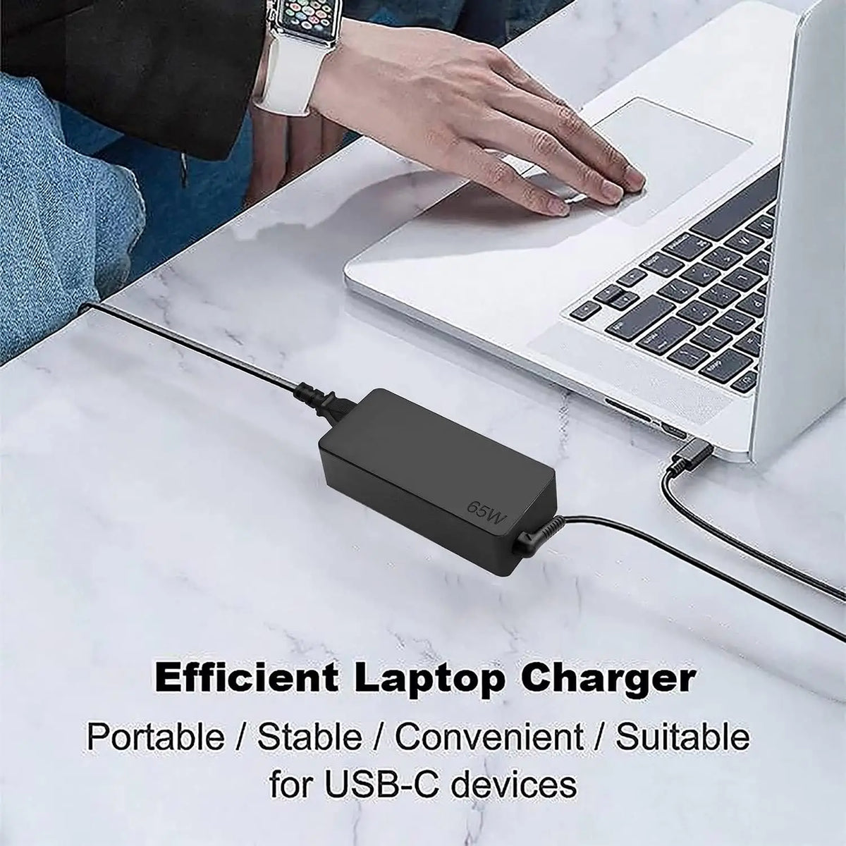 A close-up of a black 65W USB-C laptop charger plugged into a white laptop on a marble surface.