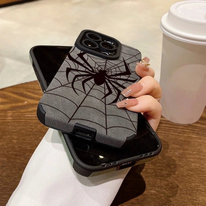 A hand holds a black phone case featuring a gray spider web design, resting on a wooden surface next to a white cup.