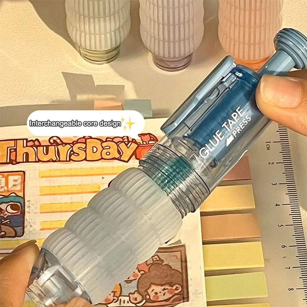 A hand holds a blue glue tape press pen with an interchangeable core design next to a ruler and colorful paper.