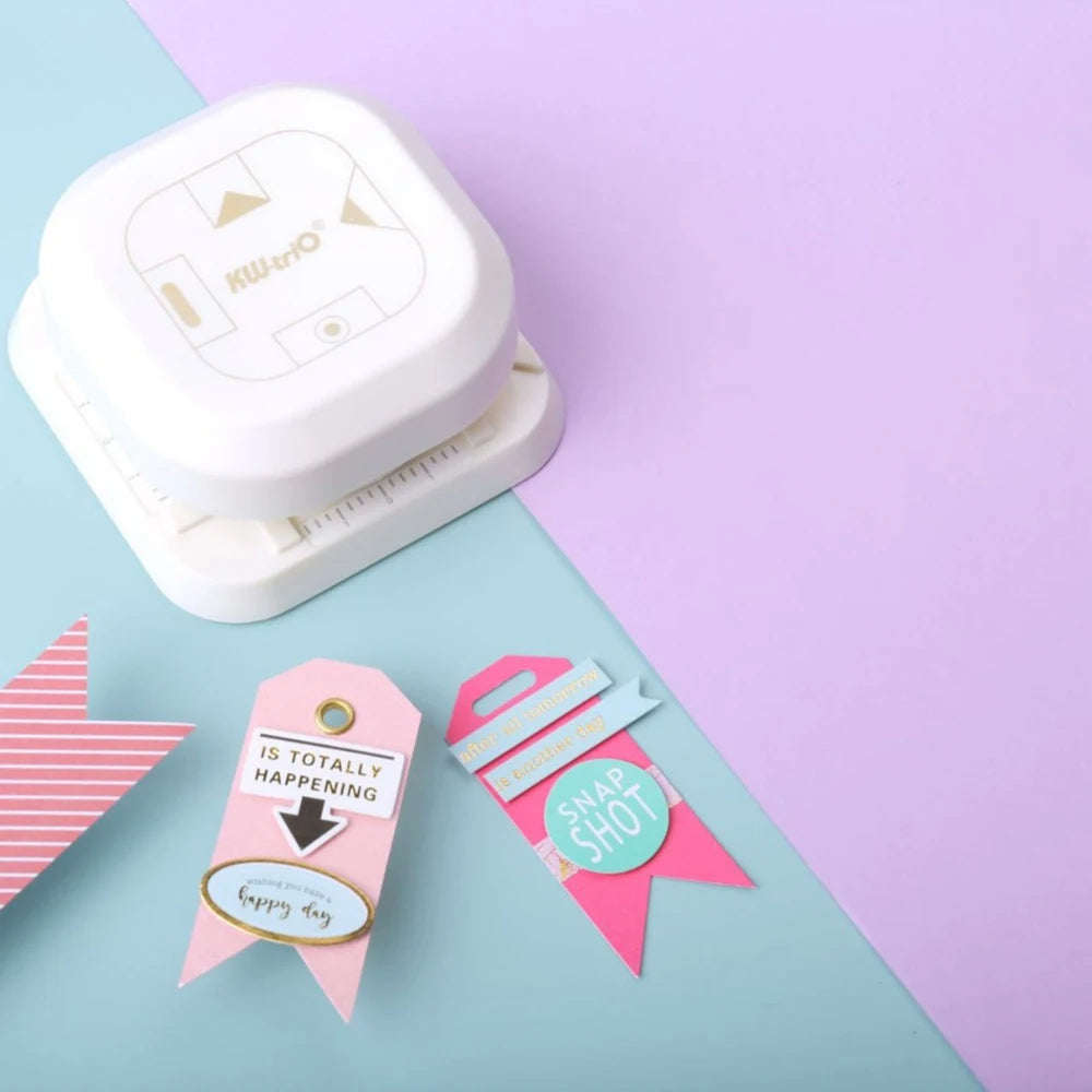 A white KW- Trio crafting tool is shown on a light blue surface next to pink and red tag punch tools.