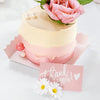 A pink and cream layered cake decorated with roses and pearl accents, presented on a pink card.