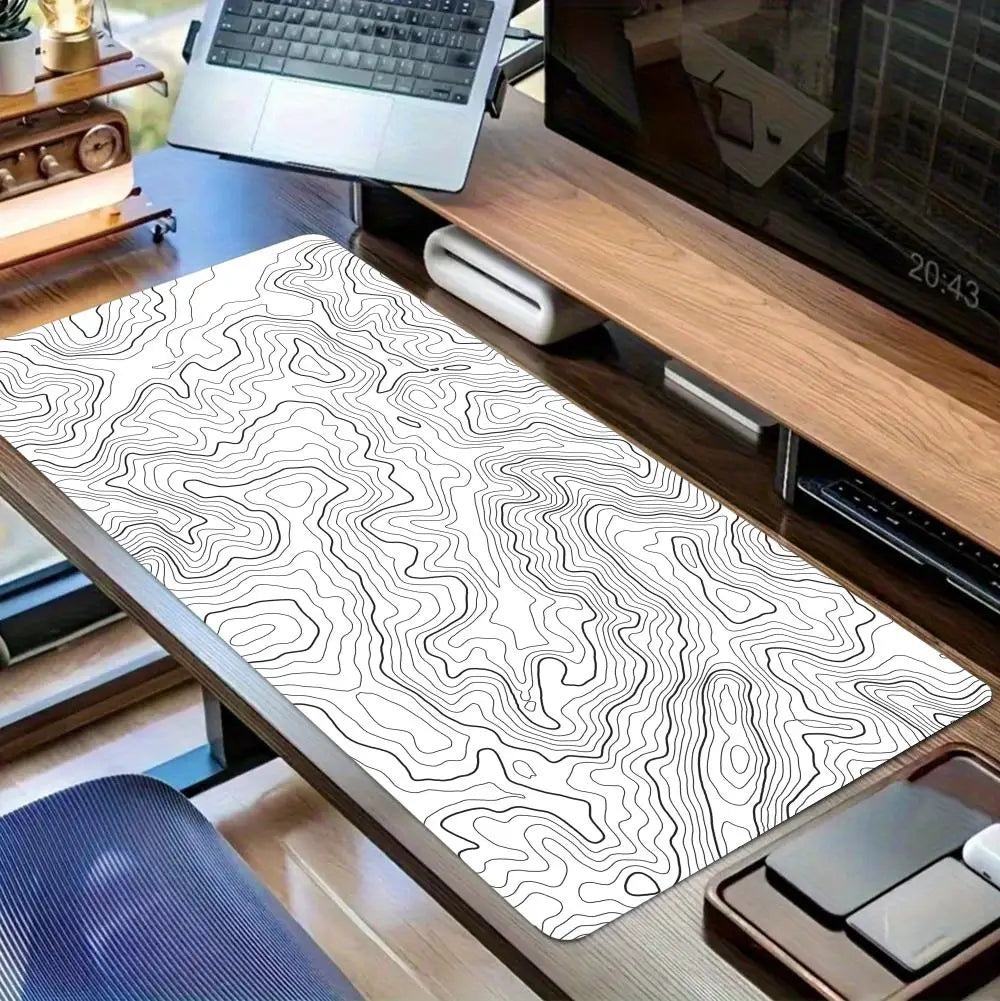 A black and white topographic map extended gaming mouse pad is displayed on a wooden desk next to a laptop.