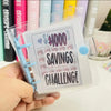 A hand holds a clear plastic binder titled Savings Challenge with a $1000 goal and savings details.