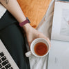 Eine Hand mit einem lila Silikon-Schnellverschluss-Armband hält eine Tasse Tee neben einem Laptop.