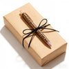 A leopard print retractable ballpoint pen with a black ribbon tied around it, resting on a kraft paper box.