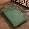 A green leather notebook with brass binding lies on a wooden surface next to a dark geometric box.