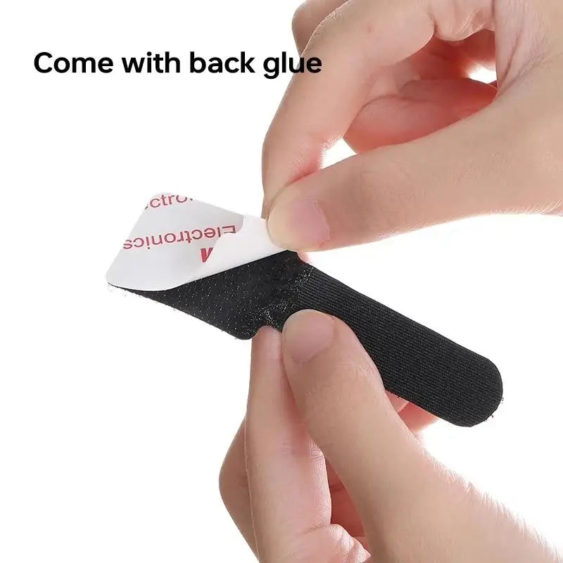 A hand is holding a black adhesive cable tie with a white label and back glue for desk organization.