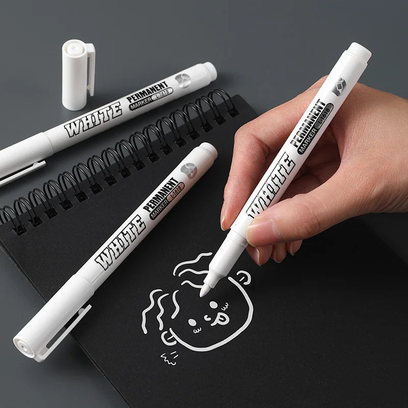 A hand holds a white permanent marker and a white cap against a black spiral notebook.
