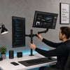 A man is adjusting a heavy duty dual monitor desk mount system with two computer monitors and a screen displaying ideas.