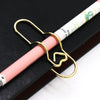 A gold heart-shaped metal pen clip is shown attached to a pink and white striped pen resting on a dark surface.