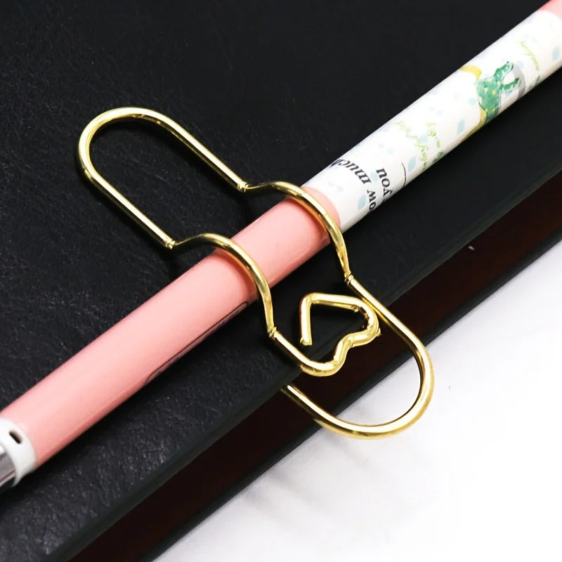 A gold heart-shaped metal pen clip is shown attached to a pink and white striped pen resting on a dark surface.