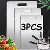 A set of three stainless steel double-sided cutting boards displayed against a dark background with fresh produce.