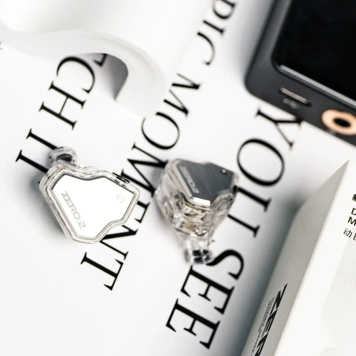 A close-up of a silver studio-grade wired in-ear monitor resting on white paper next to a smartphone.