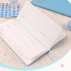 A light blue slim weekly planner journal open next to a matching blue cover and a glass.