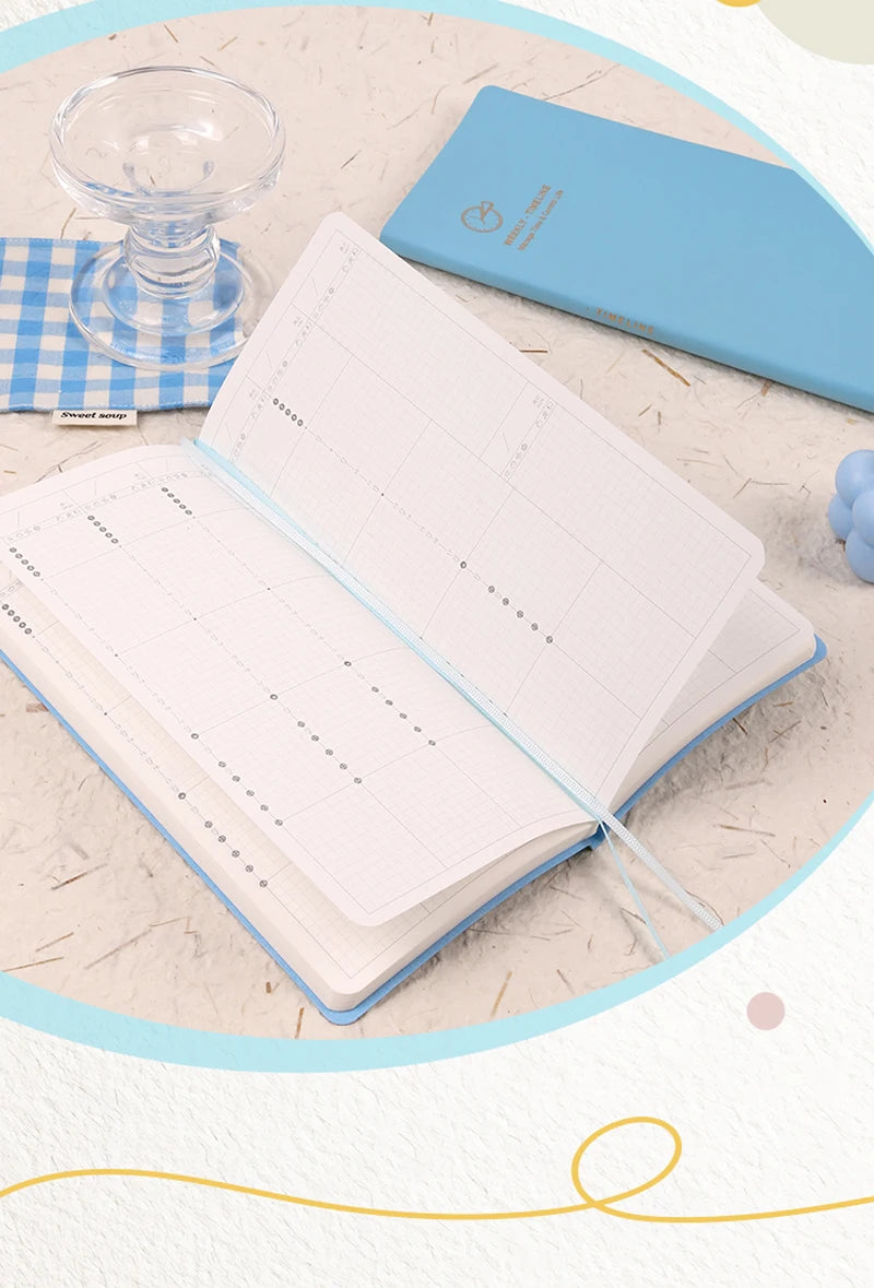 A light blue slim weekly planner journal open next to a matching blue cover and a glass.