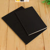 A black A6 blank sketchbook lying on a light wooden surface.