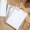 A stack of weekly meal planner and grocery list pads with various colored pages and a gold-framed watch in the background.
