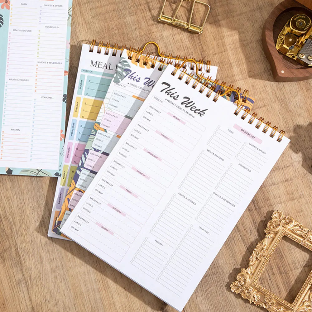A stack of weekly meal planner and grocery list pads with various colored pages and a gold-framed watch in the background.
