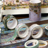A collection of decorative washi tapes in various colors and patterns laid out on a blue fabric next to an oil painting print