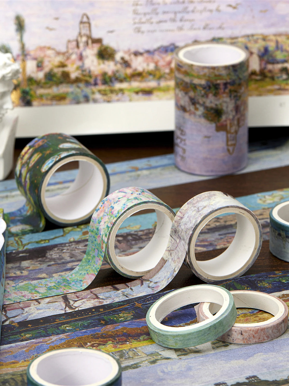 A collection of decorative washi tapes in various colors and patterns laid out on a blue fabric next to an oil painting print