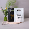 Two black and white thank you gift bags with handles are displayed next to a vase of greenery.