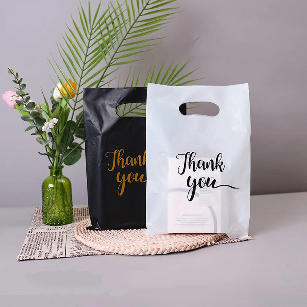 Two black and white thank you gift bags with handles are displayed next to a vase of greenery.