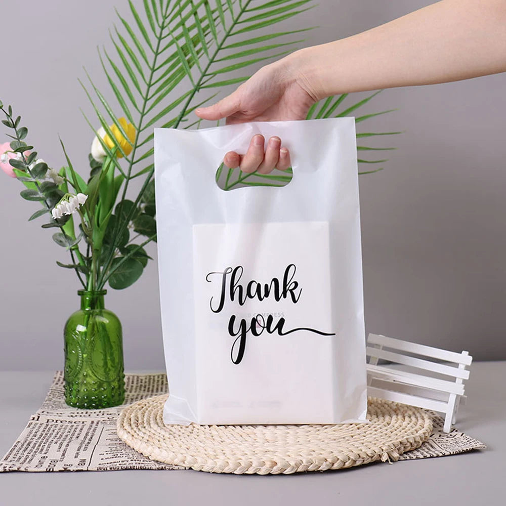 A hand holds a white thank you gift bag with a handle against a background of green palm leaves.