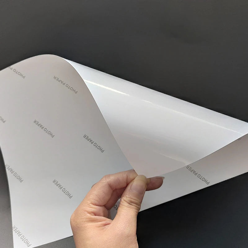 A hand is holding a sheet of white high glossy self-adhesive photo paper, showing its texture and thickness.