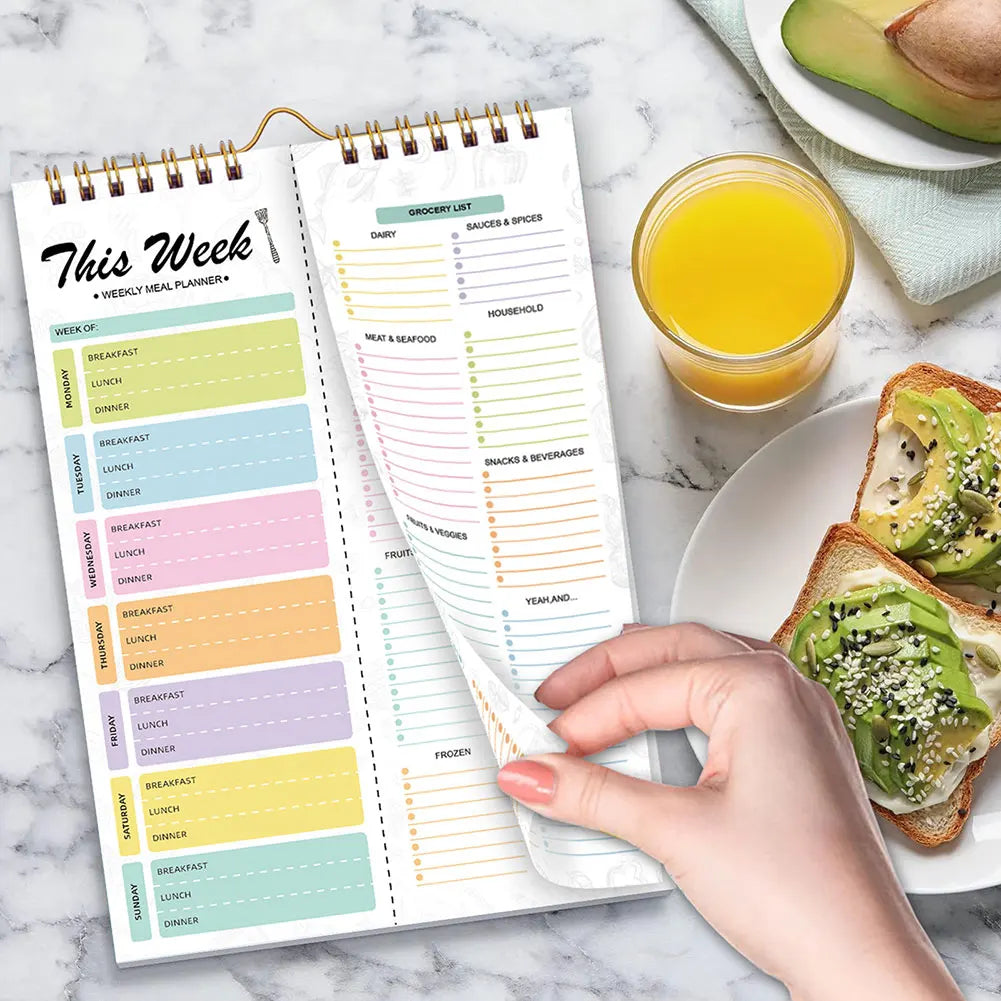 A hand holds an open weekly meal planner and grocery list pad with organized meal plans and shopping lists.