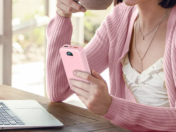 A person holds a pink power bank with a built-in AC plug and dual cables while sitting at a wooden table.