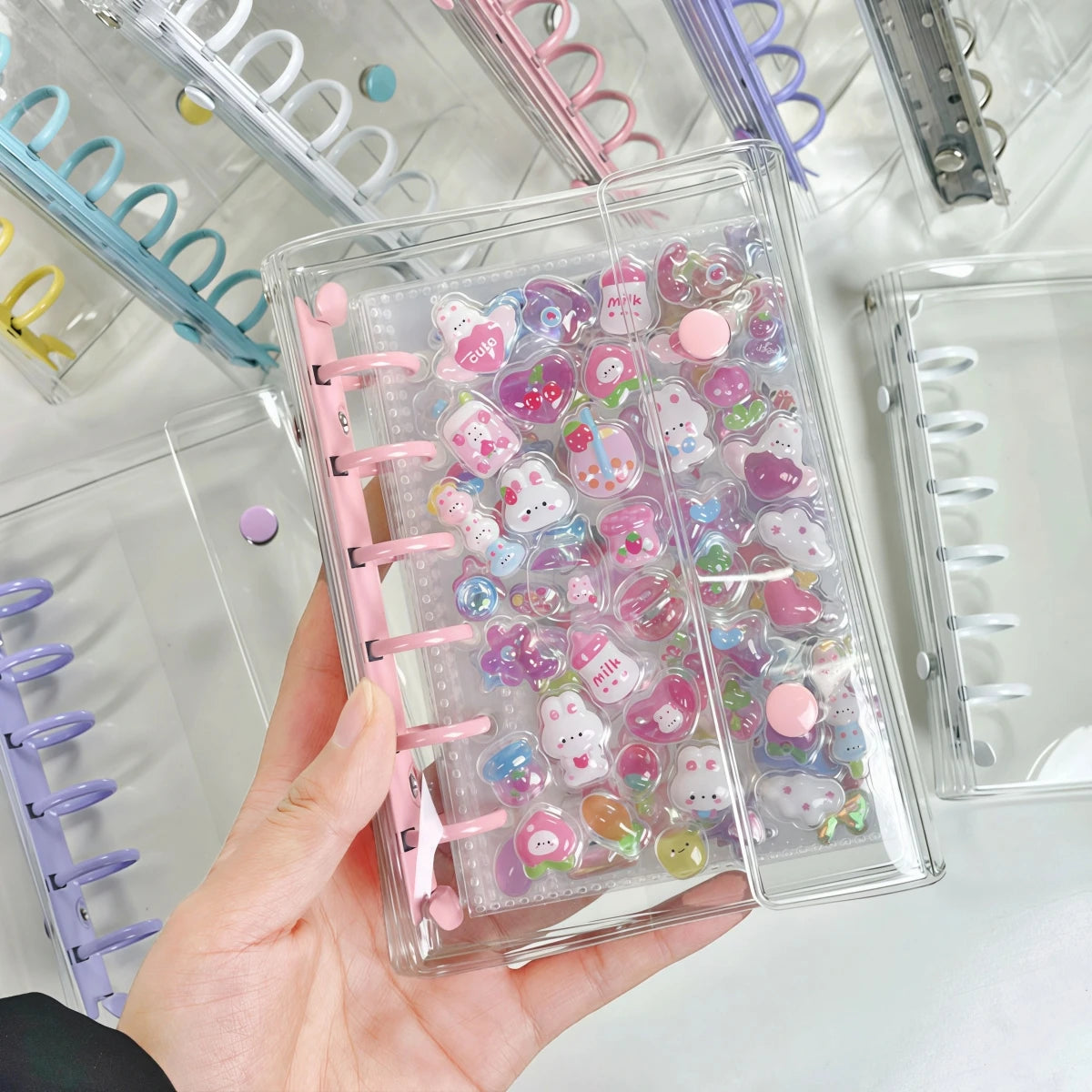A hand holds a clear double-button binder filled with colorful heart-shaped stickers for journaling and crafting.