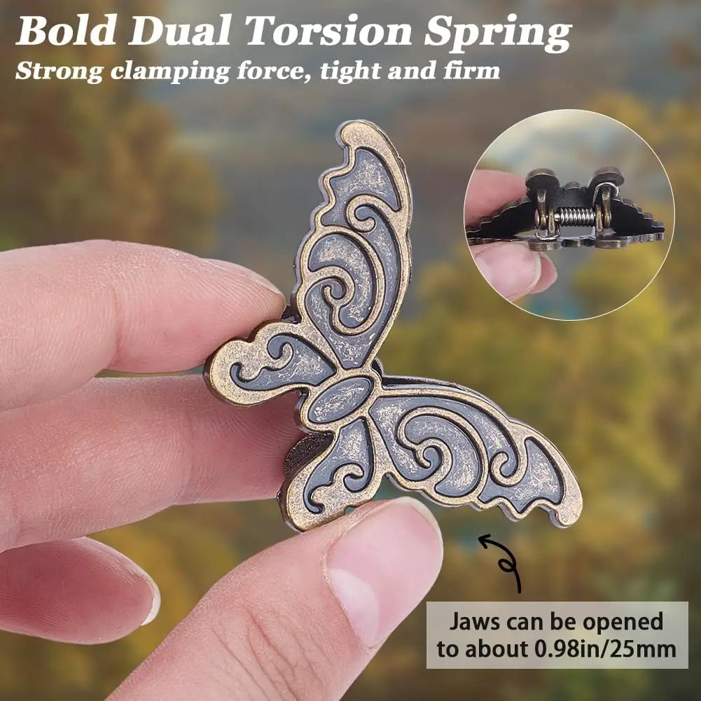 A hand holds a gold-toned butterfly shaped dual torsion spring metal book clip against a blurred background.