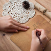 A hand is writing on a kraft paper envelope with a pen, next to a macrame coaster and a rolled parchment.