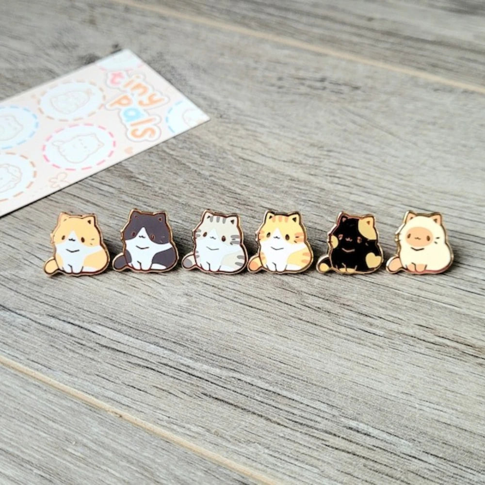 Una fila de pines de esmalte con varios gatos lindos en diferentes colores y expresiones sobre una superficie de madera.