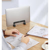 A person is using a vertical laptop stand to prop up a silver MacBook on a wooden desk while sketching.