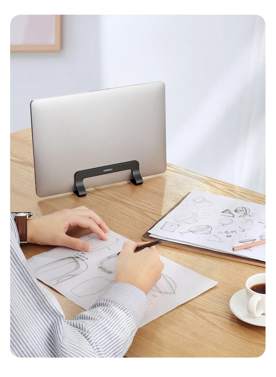 A person is using a vertical laptop stand to prop up a silver MacBook on a wooden desk while sketching.