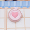 Una cinta métrica plegable en forma de corazón rosa apoyada sobre una superficie clara con un fondo de cuadrícula azul.