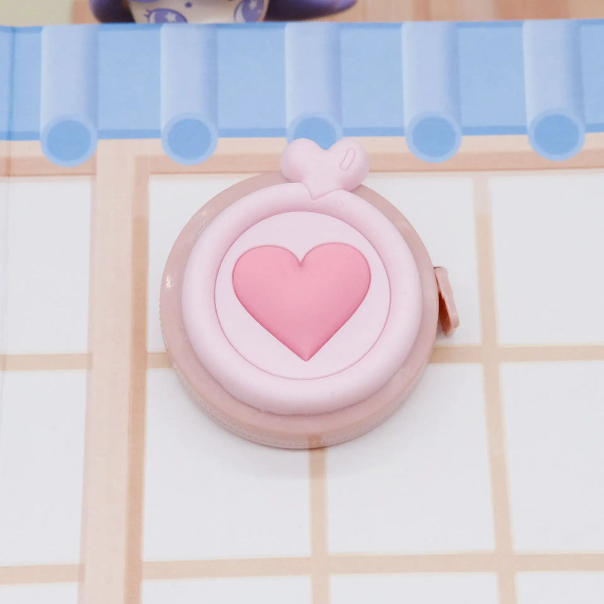 Una cinta métrica plegable en forma de corazón rosa apoyada sobre una superficie clara con un fondo de cuadrícula azul.