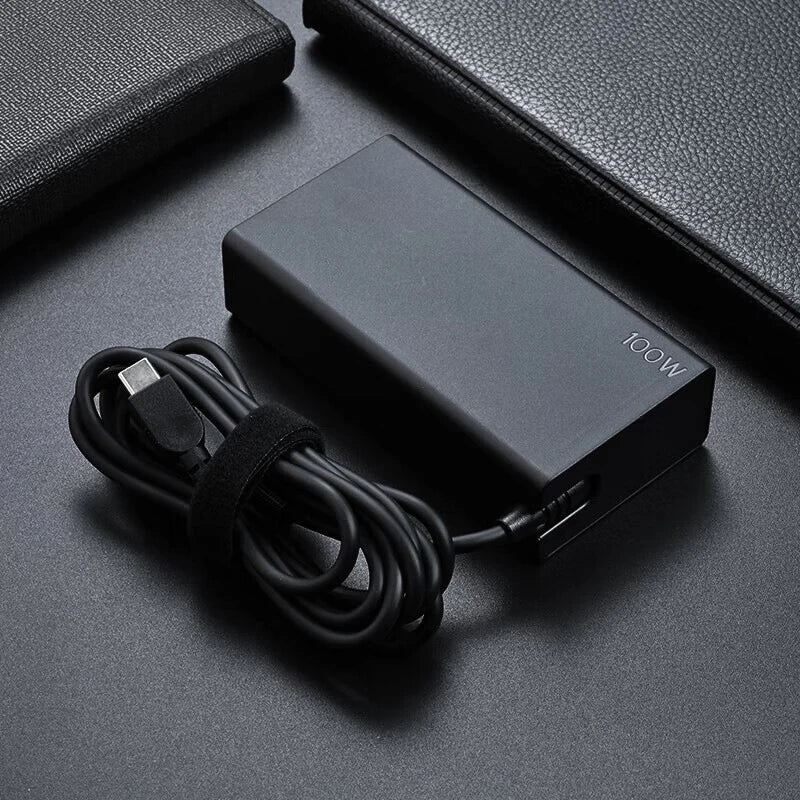 A black 100W USB-C PD laptop charger adapter with a coiled cable is shown on a dark surface.