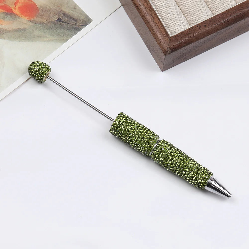 A silver pen accessory with a green rhinestone bead tip and textured body is shown on a white surface.