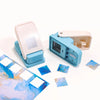 A light pink embossing punch machine with a blue housing and several small, patterned paper squares.