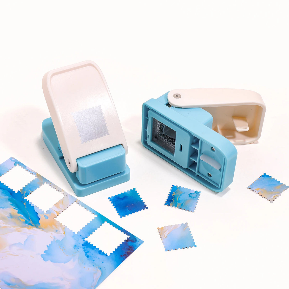 A light pink embossing punch machine with a blue housing and several small, patterned paper squares.